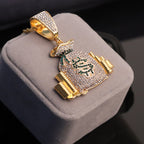 Gold-tone hip-hop money bag pendant paved with clear crystals and a green crystal dollar sign on a gray display box.