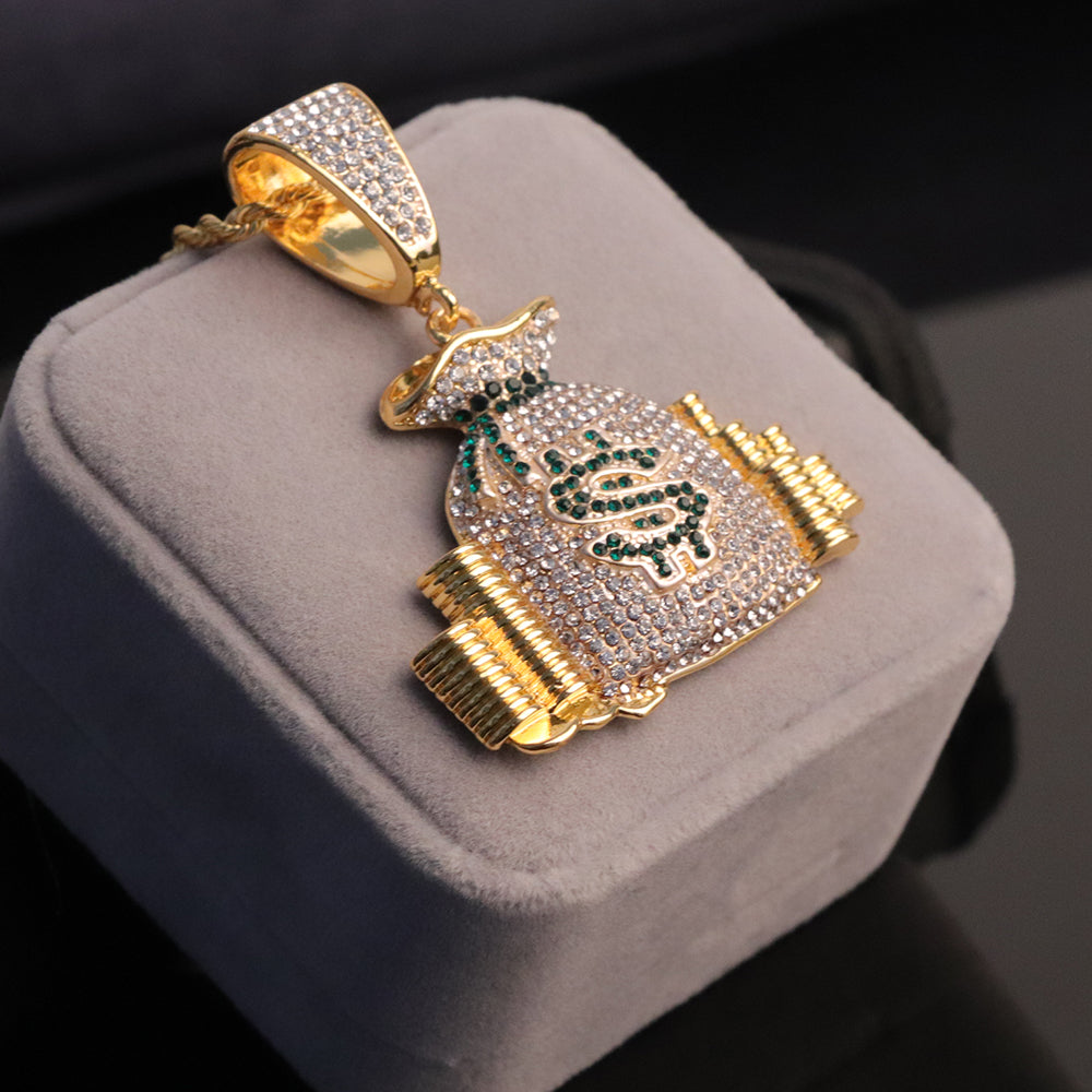 Gold-tone hip-hop money bag pendant paved with clear crystals and a green crystal dollar sign on a gray display box.