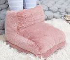 A plush, dusty rose faux fur USB foot warmer sits on a white shaggy rug with legs in light blue striped socks inside.