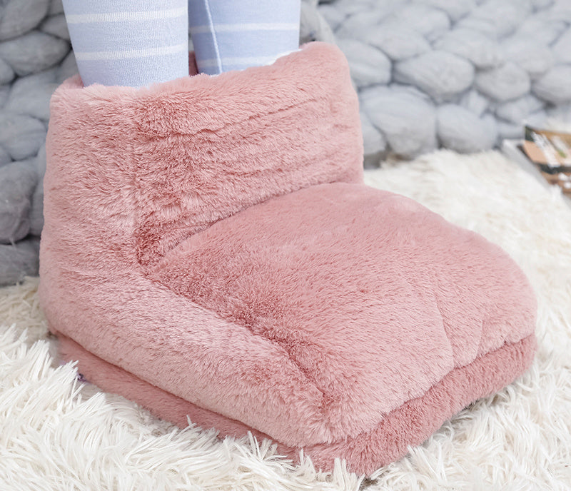 A plush, dusty rose faux fur USB foot warmer sits on a white shaggy rug with legs in light blue striped socks inside.