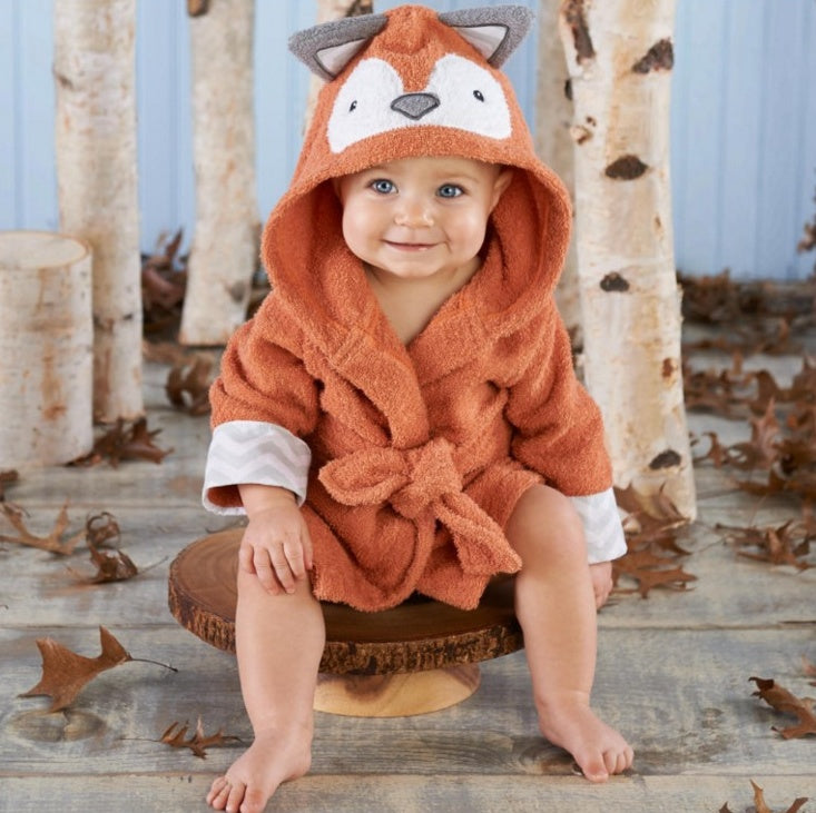 Baby wearing a soft, rust-orange cotton hooded fox bathrobe with ears and a tie-waist in a woodland setting.