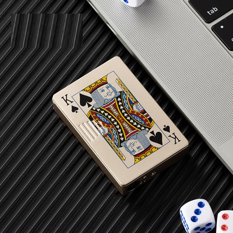 Gold metal windproof lighter featuring a King of Spades playing card design, sitting on a dark ribbed surface.