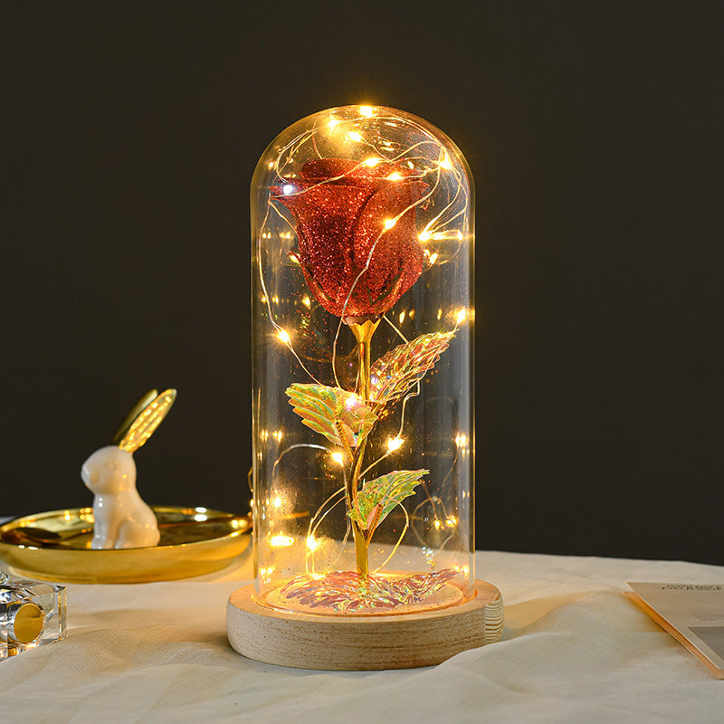 Glittery red rose inside a glass dome on a wooden base, illuminated by warm LED fairy lights.