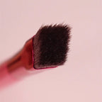 Flat-tipped brow brush with dense dark bristles and a metallic red ferrule against a soft pink background.