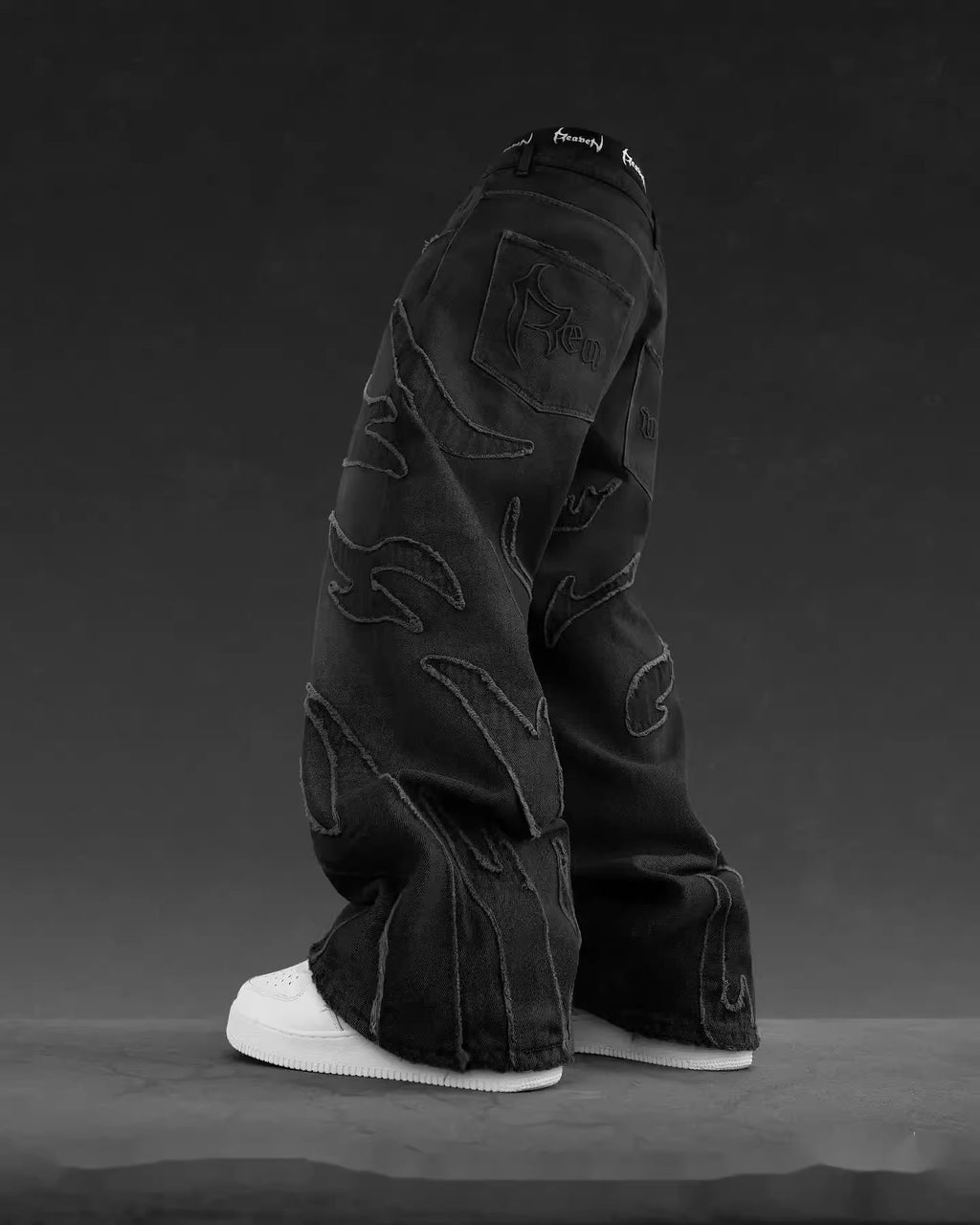 Black baggy denim jeans with flame-shaped patchwork, frayed edges, and white sneakers against a dark gray background.