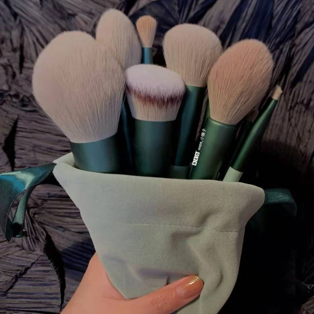 A hand holds 13 makeup brushes with forest green handles and soft, beige synthetic bristles in a sage drawstring bag.