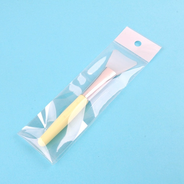Silicone facial mask brush with a yellow handle, rose gold ferrule, and white head, packaged in a clear plastic sleeve.