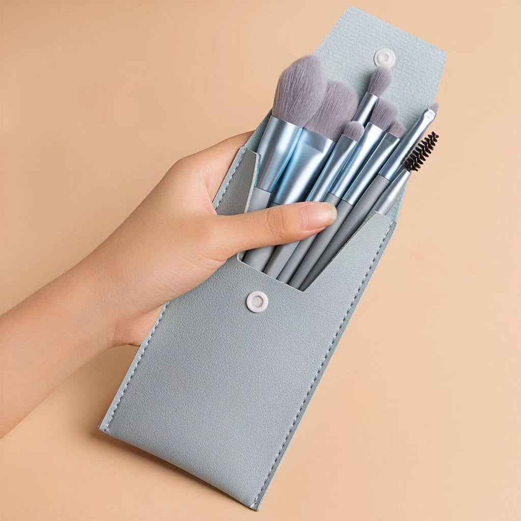 A hand holds a portable set of eight grey makeup brushes in a matching light grey vegan leather snap-button travel case.