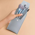 A hand holds a portable set of eight grey makeup brushes in a matching light grey vegan leather snap-button travel case.
