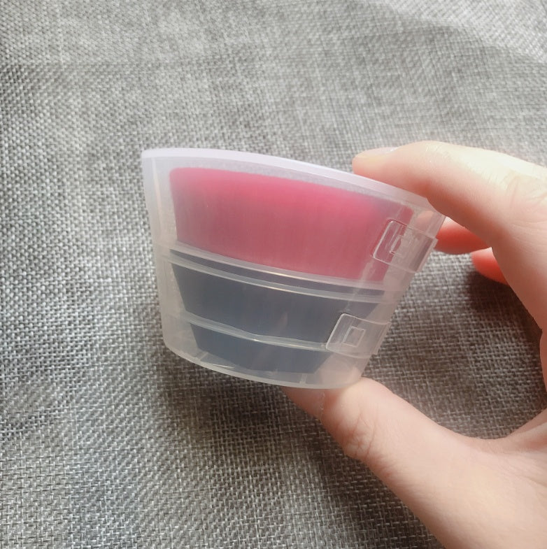 Hand holds a flat-top makeup brush with pink bristles and a black handle, encased in a clear plastic protective cover.