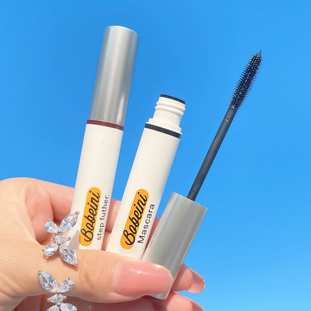 Hand holding two white Bobeini mascara tubes with silver caps and a black silicone wand against a blue sky background.