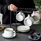 Set of white matte ceramic coffee cups with gold rims, hanging on a sleek black metal rack with matching saucers.