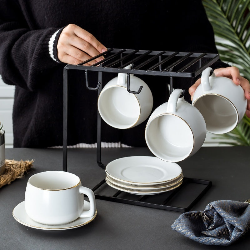 Set of white matte ceramic coffee cups with gold rims, hanging on a sleek black metal rack with matching saucers.