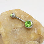Titanium steel navel barbell with two lime green faceted crystals, resting on a textured, light-colored stone.