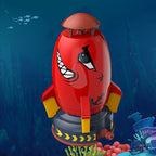 Red plastic water rocket sprinkler toy with shark-face design, yellow fins, and hose connector on a blue sea background.