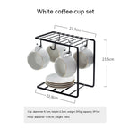 Four white matte ceramic coffee cups with gold rims hanging on a black wire rack above a stack of matching saucers.