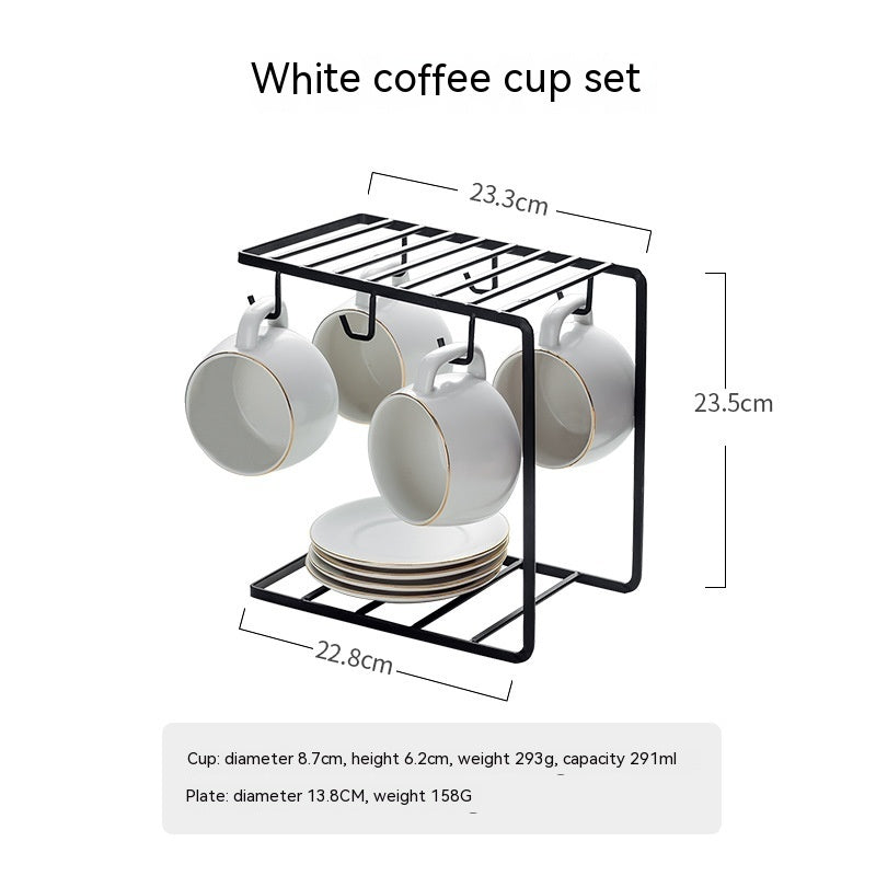 Four white matte ceramic coffee cups with gold rims hanging on a black wire rack above a stack of matching saucers.