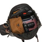 Open vintage brown leather duffle bag packed with a sweater, DSLR camera, wallet, and shoes against a white background.