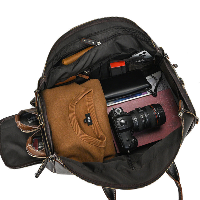 Open vintage brown leather duffle bag packed with a sweater, DSLR camera, wallet, and shoes against a white background.