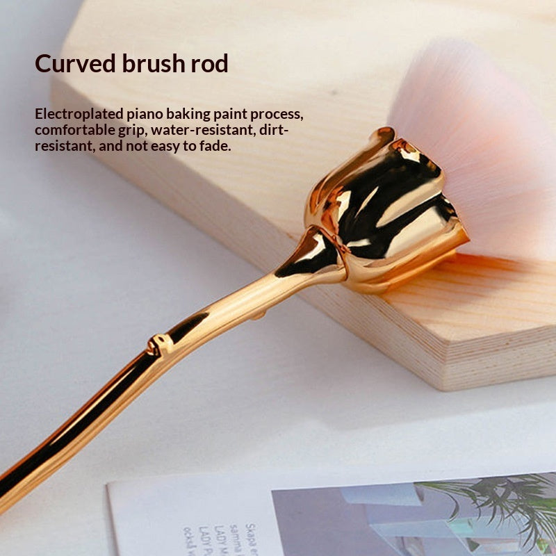 Makeup brush with soft pink bristles and a metallic gold, rose-shaped handle on a light wood surface.