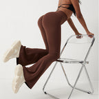 Brown high-waisted, hip-lifting flared fitness pants on a model leaning against a chair. Smooth, stretchable material.