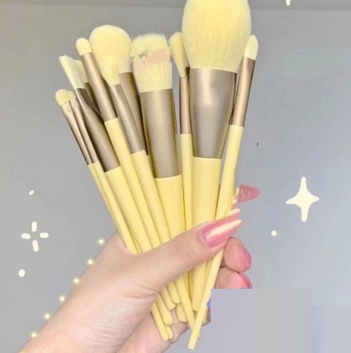 A hand holds 13 pastel yellow makeup brushes with matte metallic ferrules against a light background with sparkle icons.