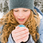 A woman in winter gear holds a round, metallic blue portable hand warmer to her face in a snowy outdoor setting.