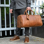 A person holding a tan leather vintage-style travel duffle bag with sturdy top handles and metal base studs.