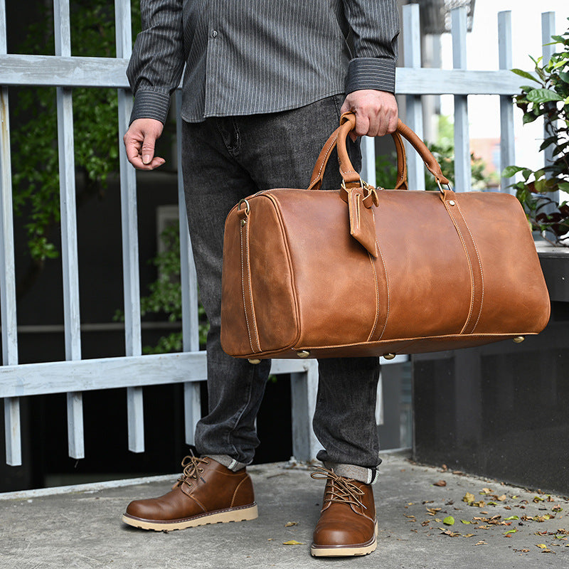 A person holding a tan leather vintage-style travel duffle bag with sturdy top handles and metal base studs.