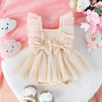 Beige baby jumpsuit with a ruffled tulle skirt, two front bows, and soft fabric, displayed with flowers and decor.