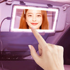 A hand touches a rectangular LED makeup mirror attached to a car sun visor with a bright, illuminated light frame.