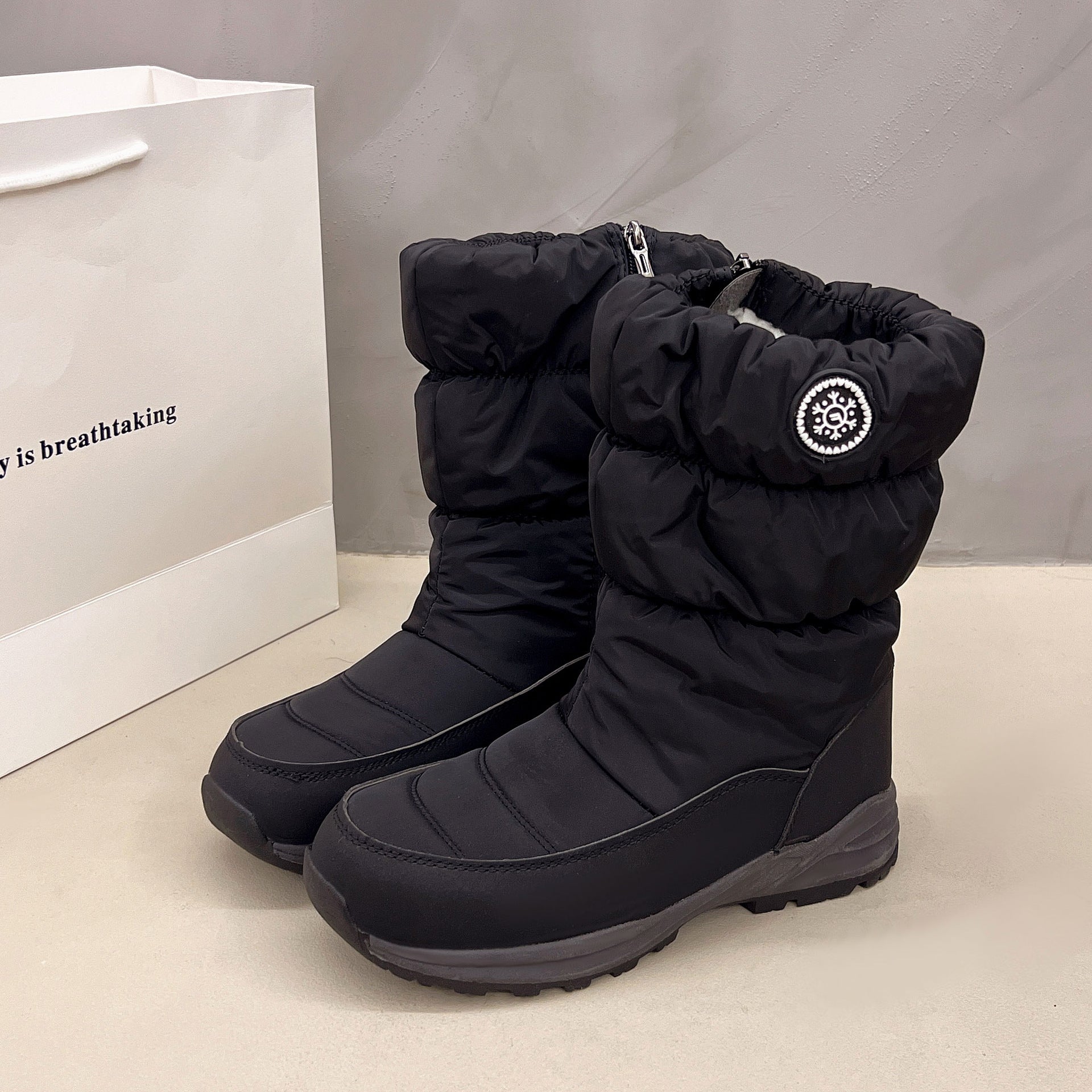 Black mid-calf snow boots with quilted fabric, a fuzzy lining, side zipper, and a circular snowflake emblem patch.