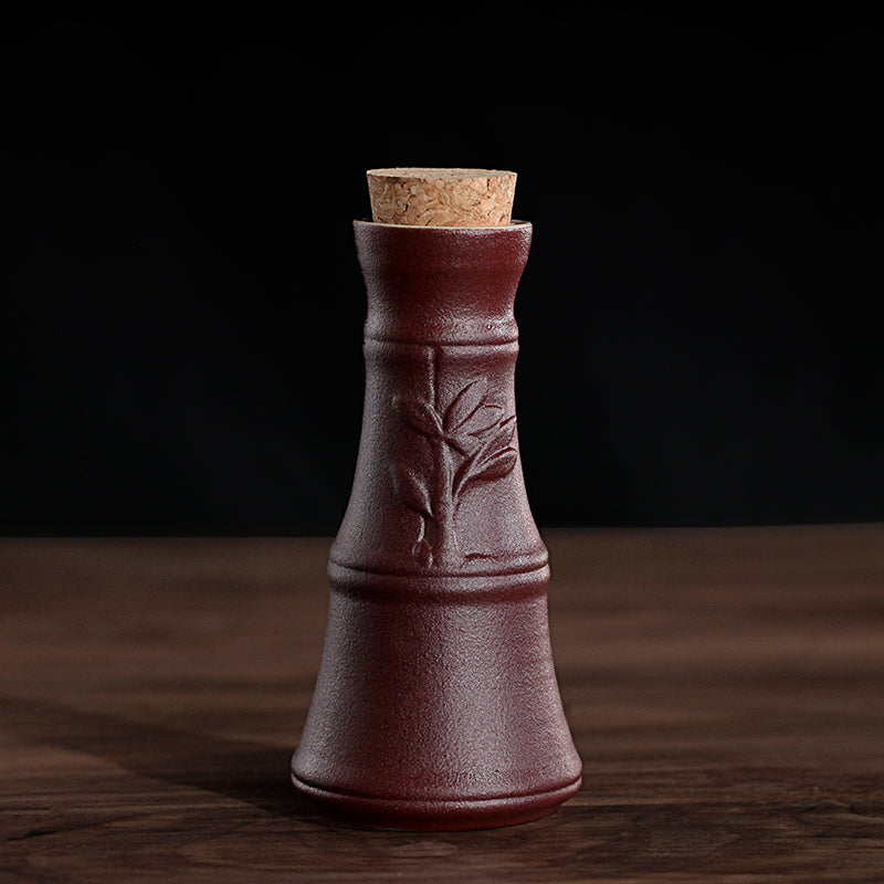 A rustic, dark red ceramic wine carafe featuring a bamboo relief pattern and a natural cork stopper on a wooden surface.