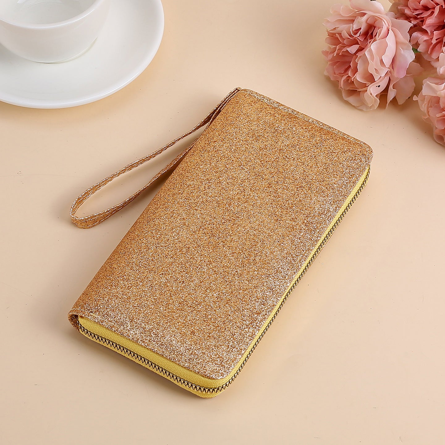 Gold glittery zip-around women's clutch with a matching wrist strap, set on a beige surface with floral accents.