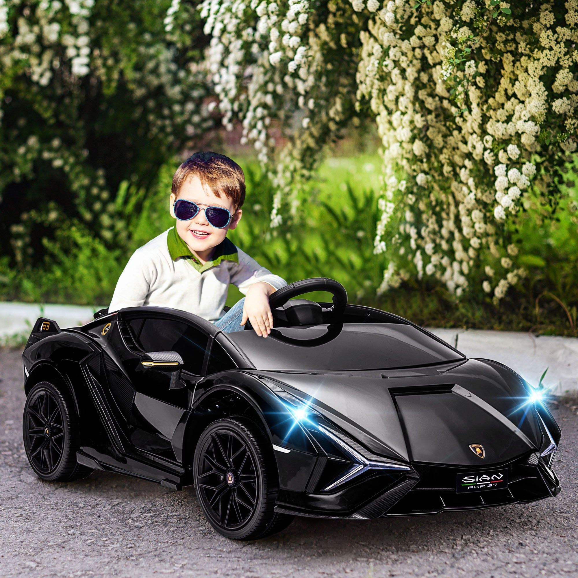 A young boy in sunglasses sits in a black 12V Lamborghini Sian electric toy car with working LED headlights.