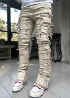 Beige stacked denim jeans featuring distressed horizontal rips with frayed edges, worn with white and red sneakers.