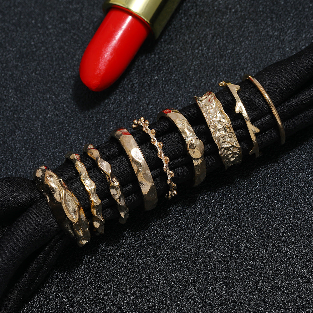 A set of eight gold-tone metal rings with varied textured, organic, and minimalist designs on black fabric.