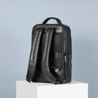 Black leather business backpack with padded straps, mesh back panel, and top handle, set on a white block against blue.