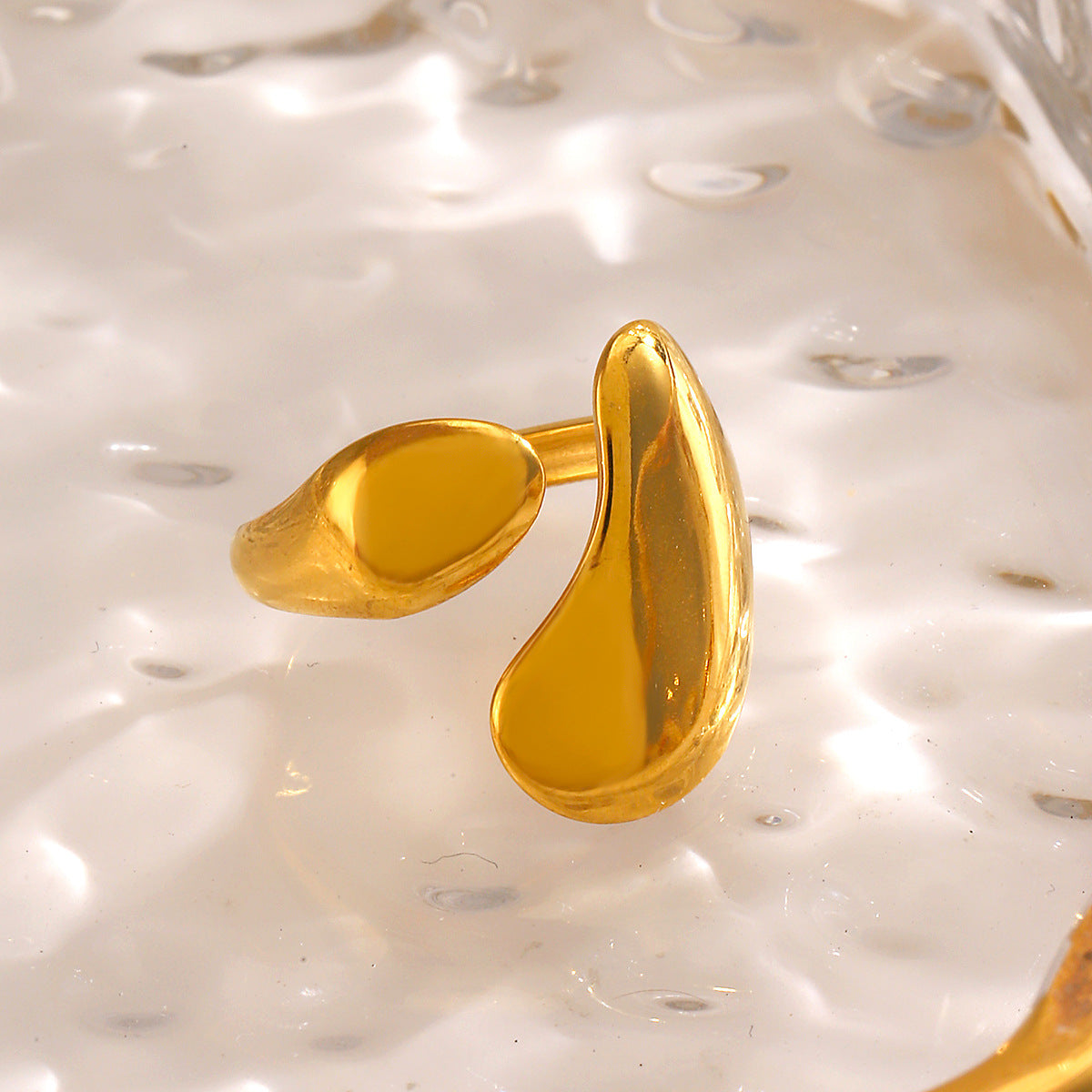 Gold-tone stainless steel ring with an open, asymmetrical organic shape, resting on a textured glass surface.