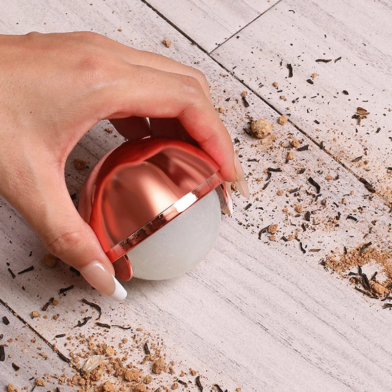 Hand holding a metallic pink, spherical, reusable gel lint roller over a wooden surface covered in loose debris.
