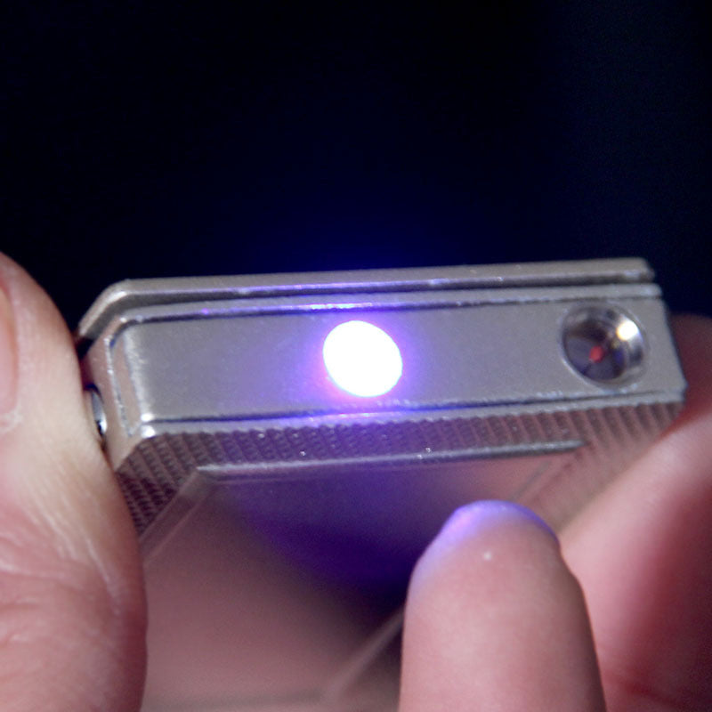 Silver metallic playing-card style lighter with a bright blue LED light feature, held between fingers.