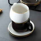 Stacked white and black matte ceramic coffee cups with gold rims, resting on matching saucers on a dark surface.