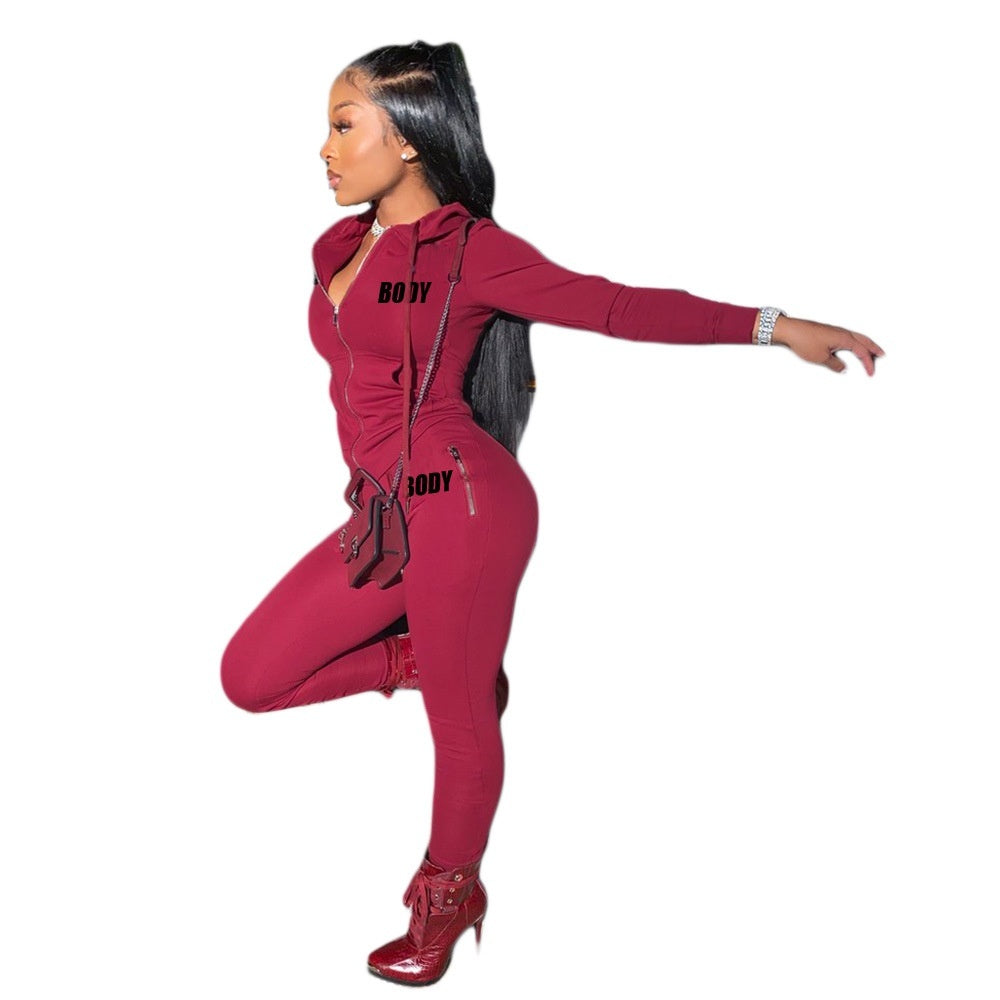 A woman in a fitted, maroon zipped hooded bodysuit with "BODY" print, styled with a crossbody bag and red heels.