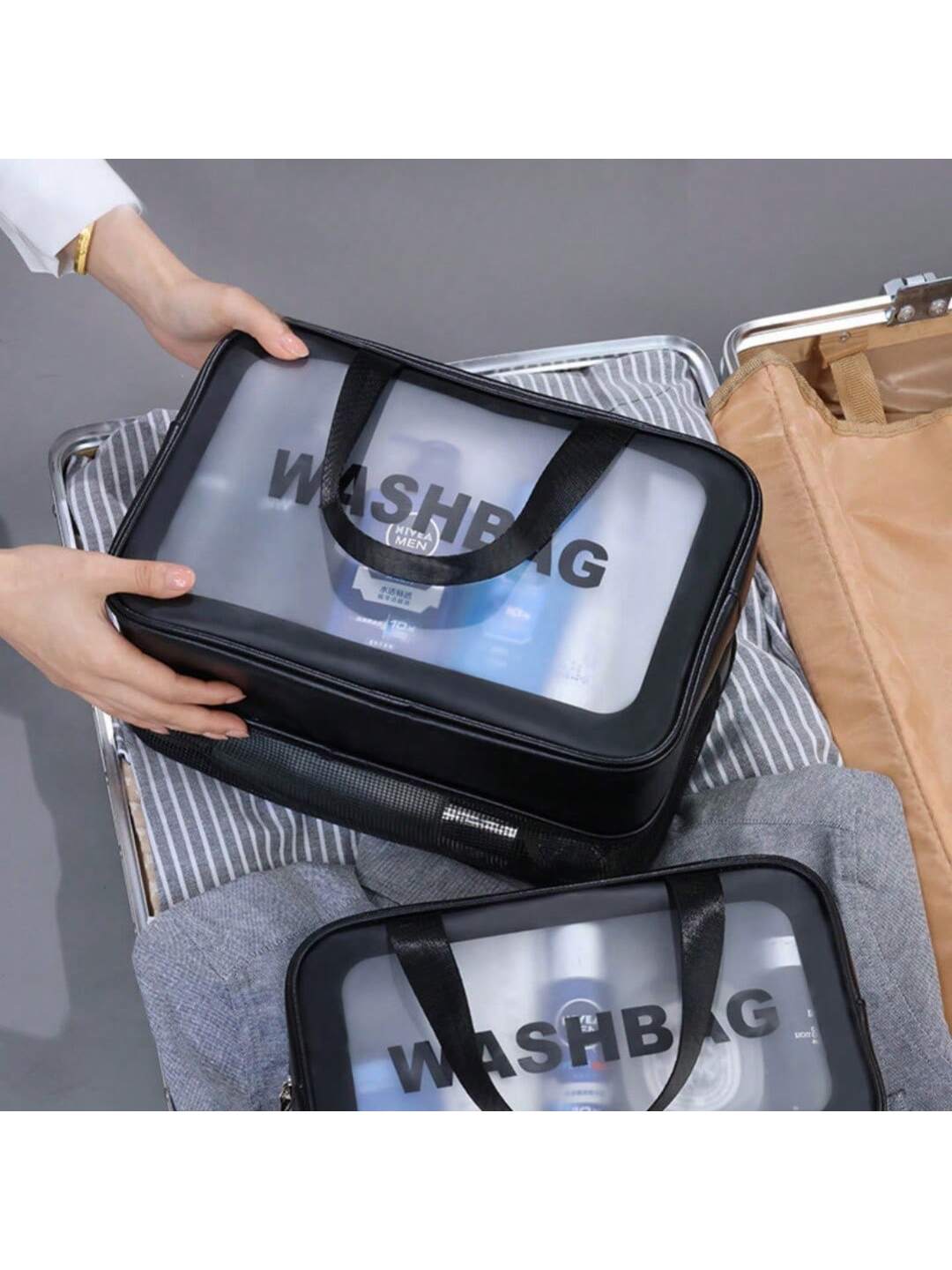 Hands holding a black transparent PVC travel washbag with "WASHBAG" printed on it, featuring black fabric handles.