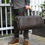 A person holds a brown, vintage-style leather duffle bag with side zippered pockets and sturdy handles.