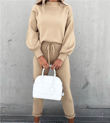 A woman wears a beige long-sleeve knit lounge set with a mock-neck top and drawstring pants, holding a white handbag.