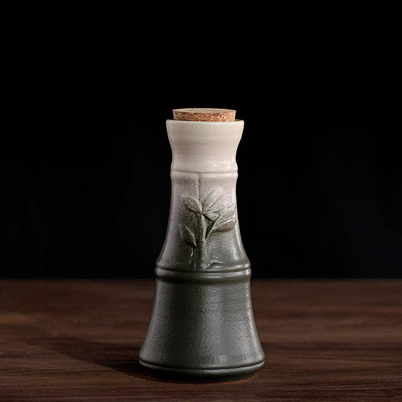 A tapered, ceramic sake pot fades from dark green to cream, featuring a leaf design and a small cork stopper.