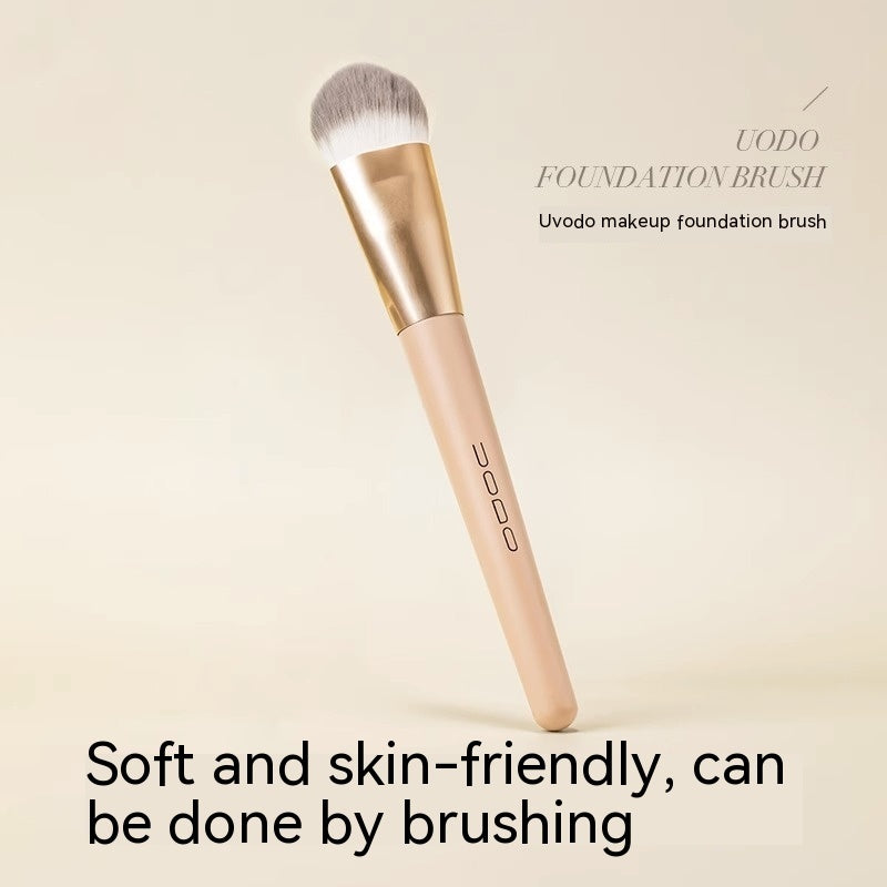 Uodo foundation brush with a flat, soft grey-white head, gold ferrule, and a beige handle on a neutral background.