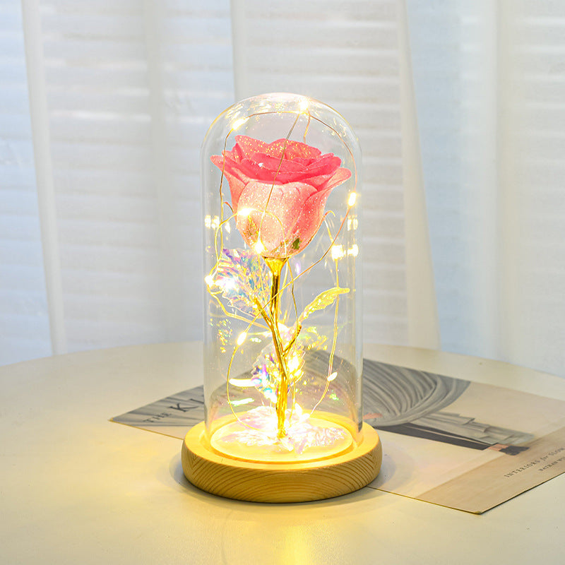 Pink glitter rose with a gold stem inside a glass dome on a wooden base, illuminated by warm LED fairy lights.