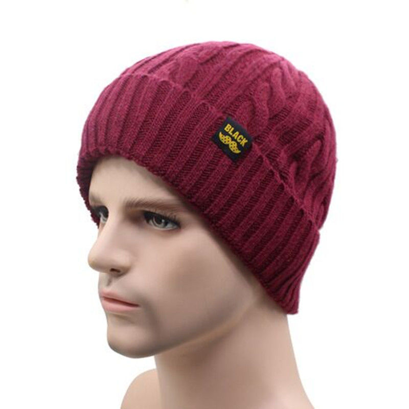 A man wearing a burgundy, cable-knit winter beanie with a ribbed cuff and a small black and yellow logo patch.